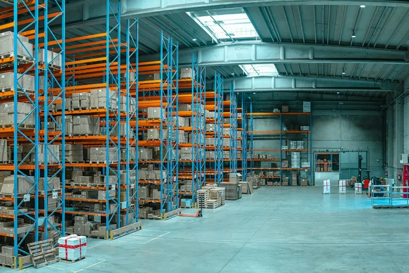 Ruime magazijnhal met blauw-oranje palletstellingen vol dozen en een vrije rijgang