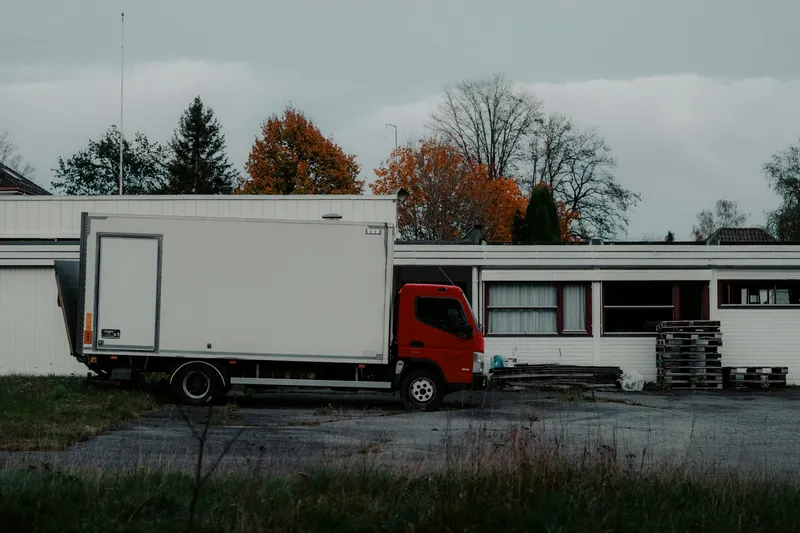 Bakwagen met witte laadbak en rode cabine bij een loods met gestapelde paletten